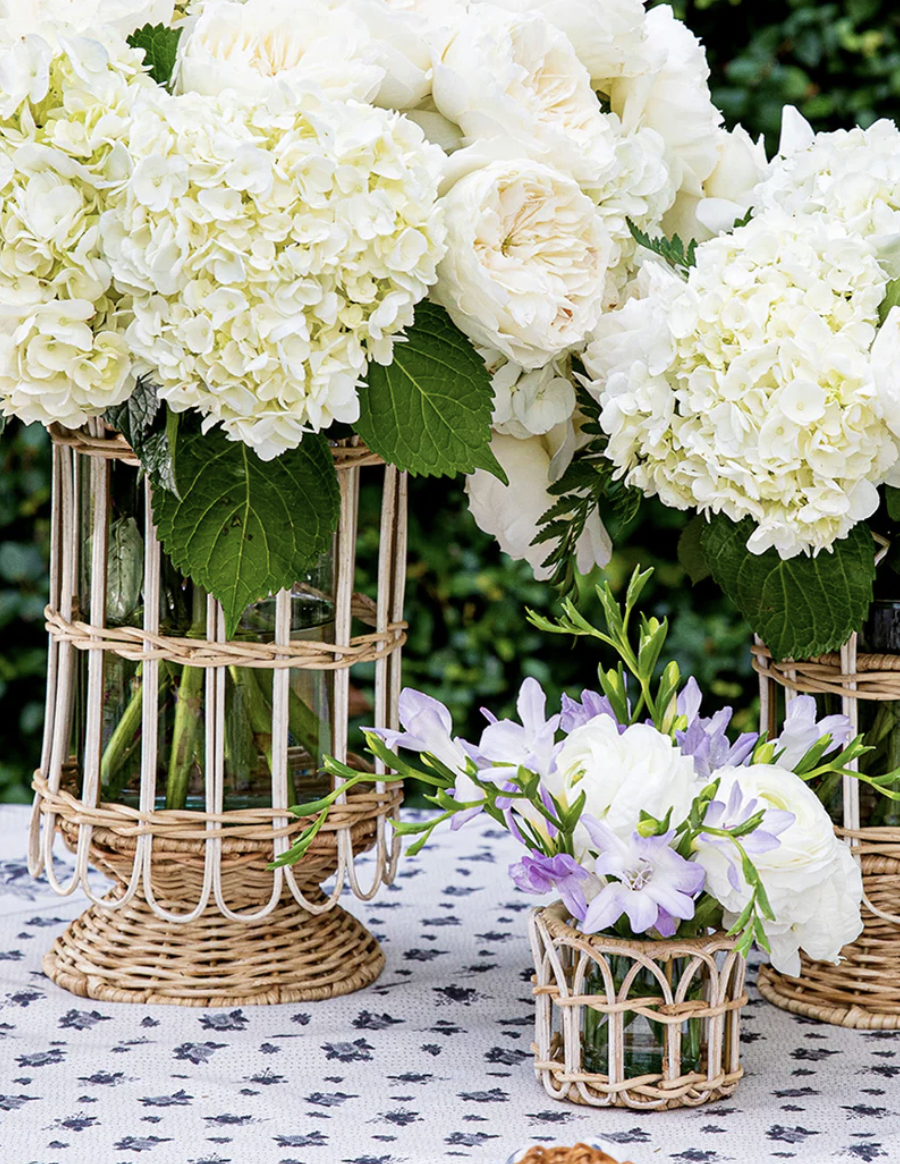 A close-up of rattan-wrapped glass vases filled with white hydrangeas, cream roses, and delicate purple blooms. The vases sit on a patterned white and navy tablecloth in an outdoor garden setting, creating a fresh, elegant floral centerpiece display.